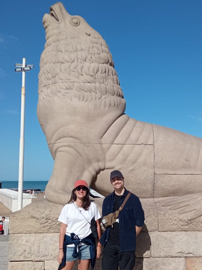 Two young people, standing in front of a Sea-Lion sculpture in sunny weather. Calm Atlantic sea in the background.